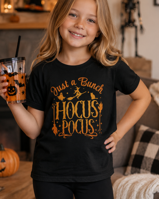 A young girl smiling in a cozy living room, wearing a black graphic t-shirt that reads 'Just a Bunch HOCUS POCUS' in orange. She is holding a clear plastic cup with a black straw and an orange pumpkin design.
