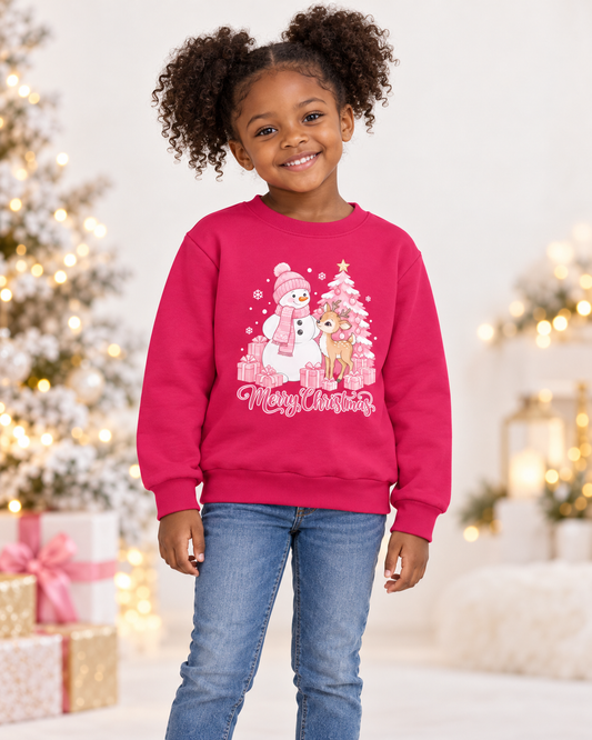 A young girl with curly hair wearing a bright pink crewneck sweatshirt with a Christmas snowman and reindeer graphic, paired with blue jeans, standing in a festive holiday setting with a decorated Christmas tree and warm lights in the background.