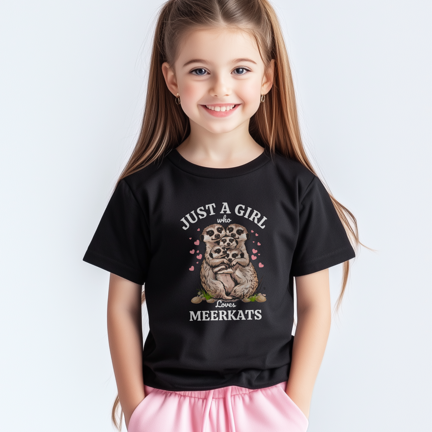 Smiling young girl wearing a black T-shirt with the text “Just a Girl Who Loves Meerkats” and a cute meerkat family graphic surrounded by pink hearts, standing against a light background.
