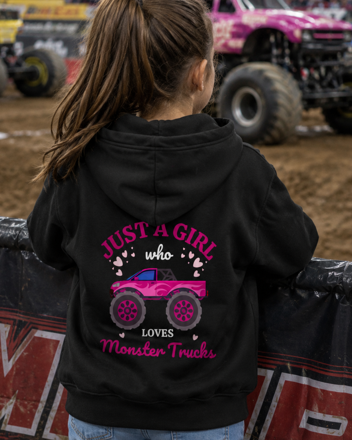 A young girl viewed from behind wearing a black hoodie with a pink monster truck graphic and the text 'JUST A GIRL who LOVES Monster Trucks'. The background shows a dirt arena with a pink monster truck and other race cars.