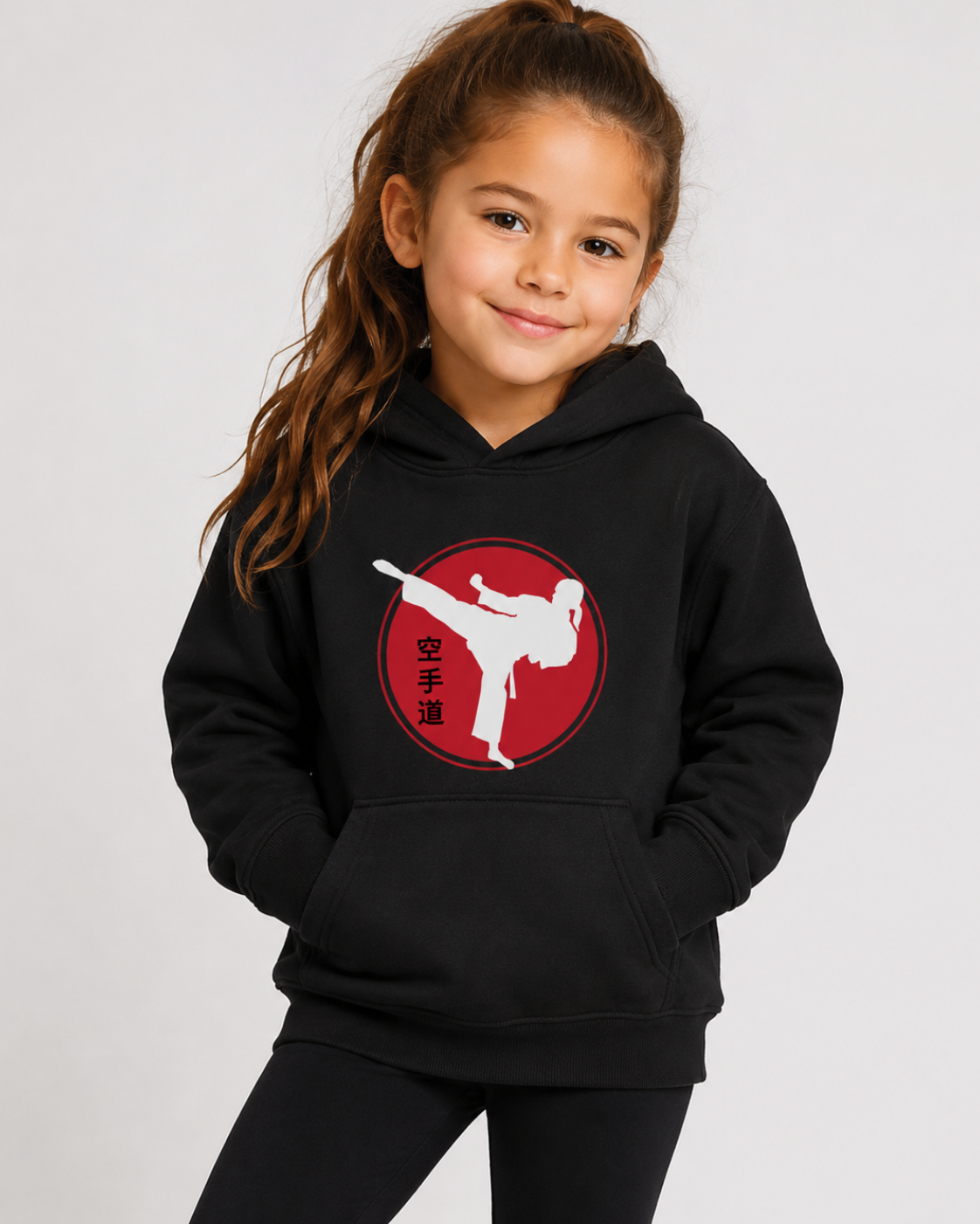 A young girl with her hair in a ponytail is wearing a black pullover hoodie with a red and white graphic on the chest depicting a martial arts kick, alongside black leggings, against a light grey studio background.