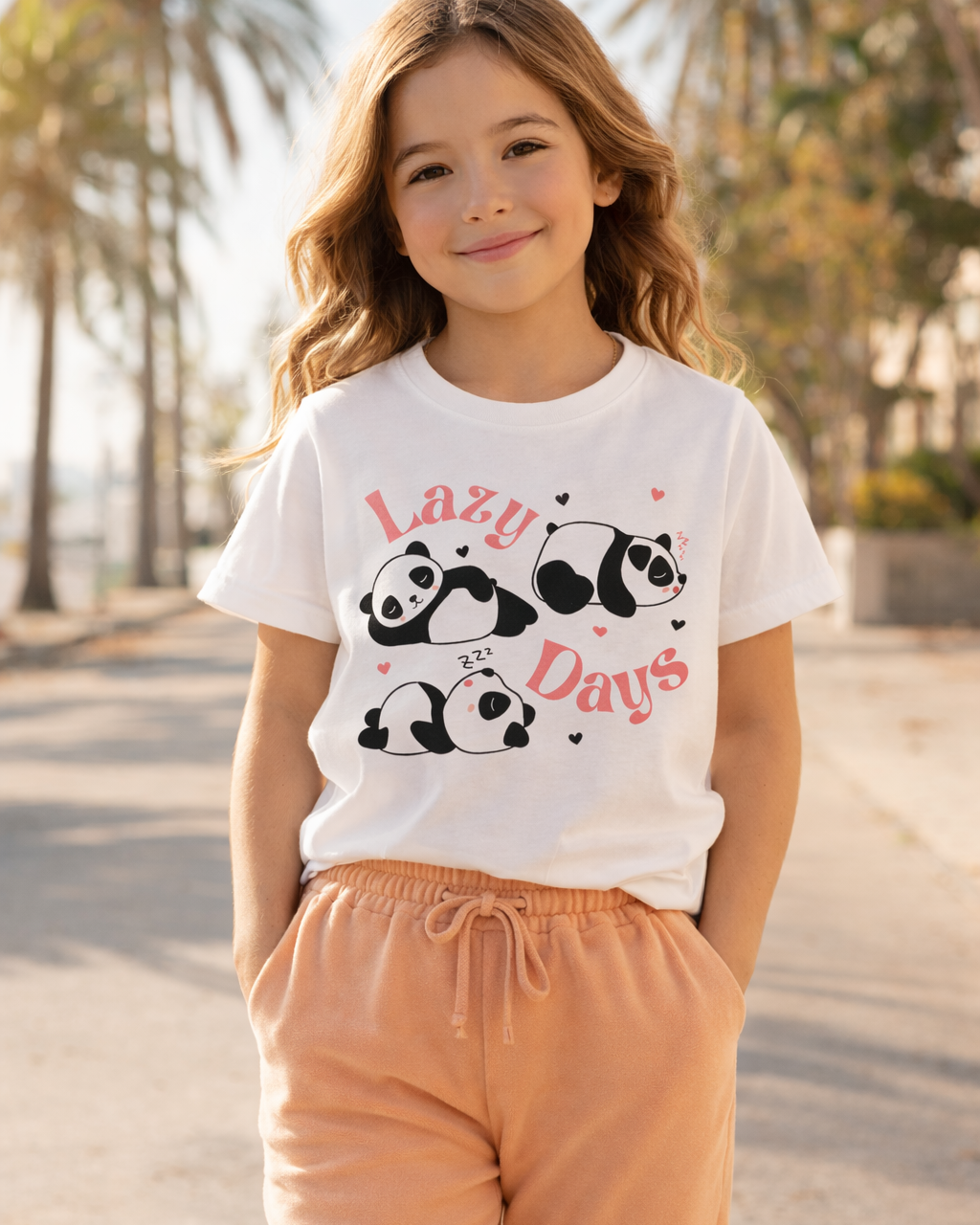 Young girl wearing a white kids “Lazy Days” panda graphic t-shirt styled with apricot joggers outdoors in warm sunlight.