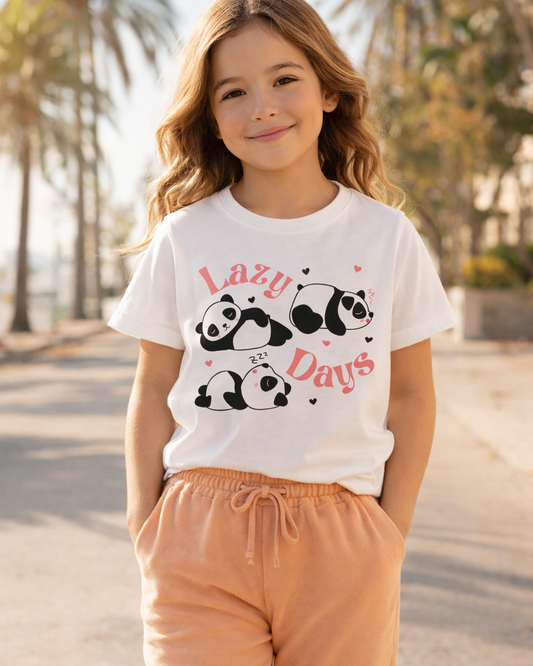 Young girl wearing a white kids “Lazy Days” panda graphic t-shirt styled with apricot joggers outdoors in warm sunlight.