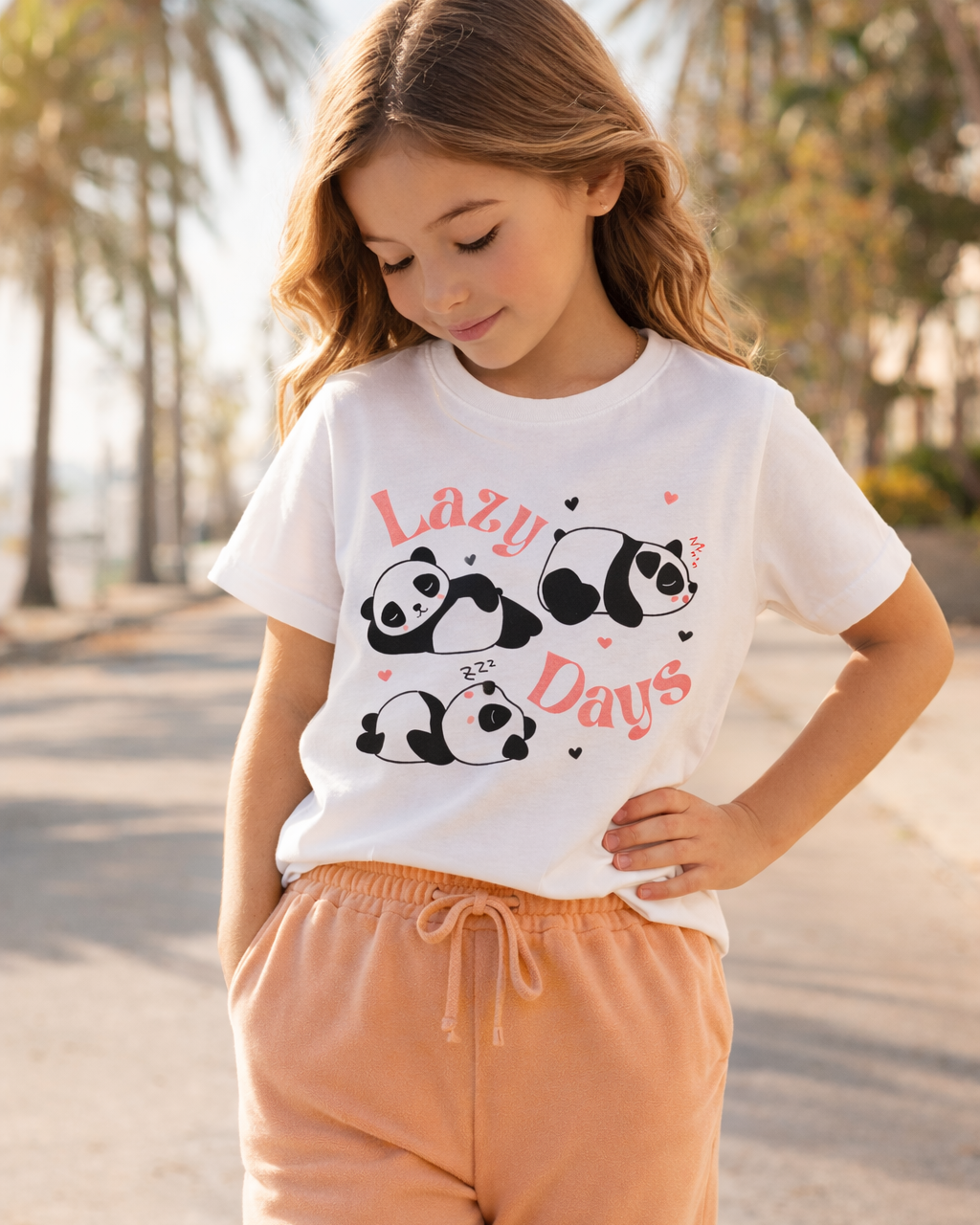 Girl modelling a white “Lazy Days” panda print kids t-shirt with apricot joggers, looking down at the design in natural outdoor light.