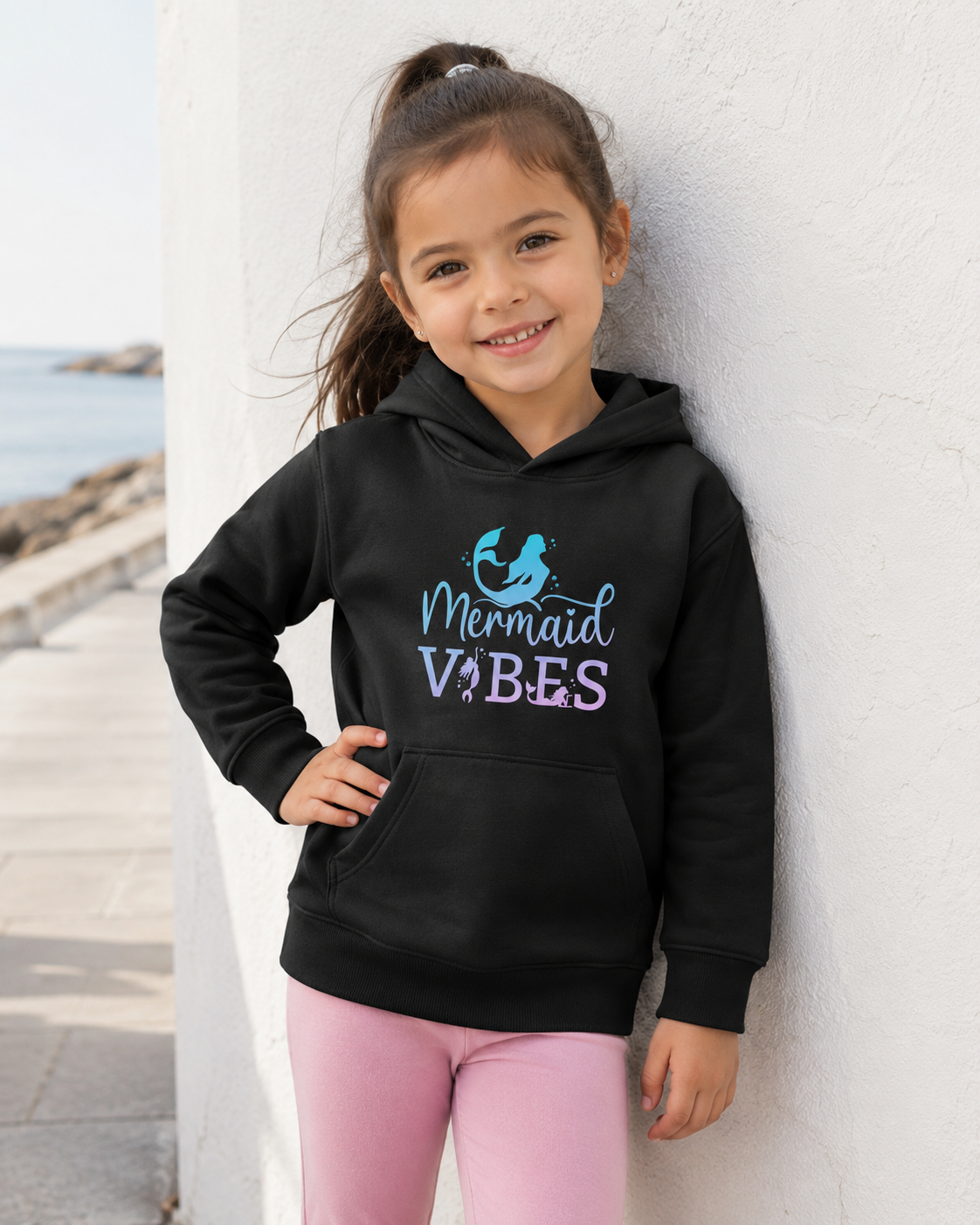 A young girl with her hair in a ponytail smiles while leaning against a white wall outdoors. She is wearing a black pullover hoodie with a 'Mermaid Vibes' graphic in blue and purple, paired with pink leggings.