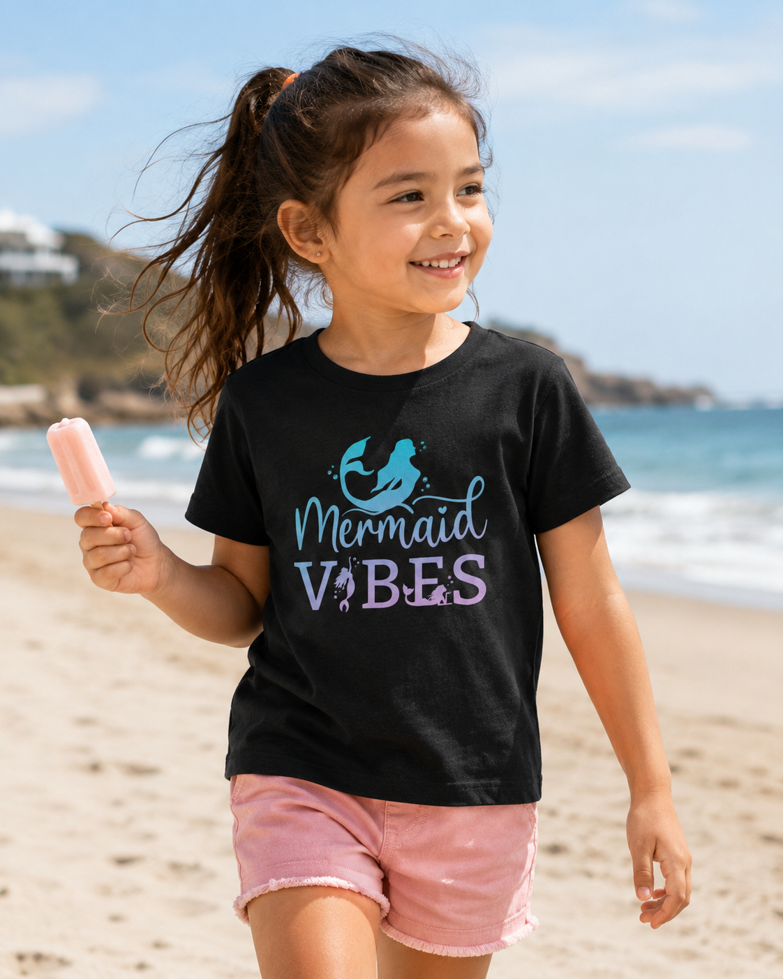 A young girl with her hair in a ponytail walks on a sandy beach wearing a black graphic t-shirt with swimming mermaids text that reads 'Mermaid VIBES' in ombre blue, pink and purple, paired with pink shorts and holding a pink ice lolly.