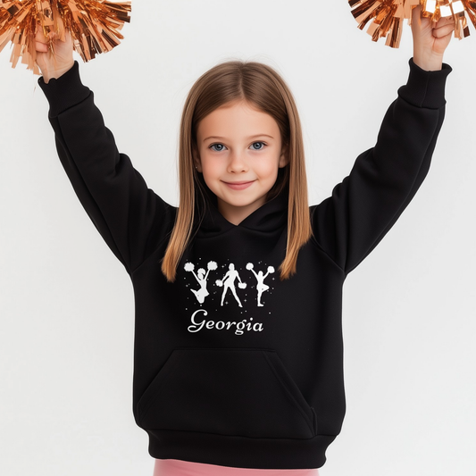 Young girl holding copper-coloured pom poms in the air while wearing a black personalised cheerleading hoodie with white cheerleader silhouettes and the name “Georgia” printed on the front.