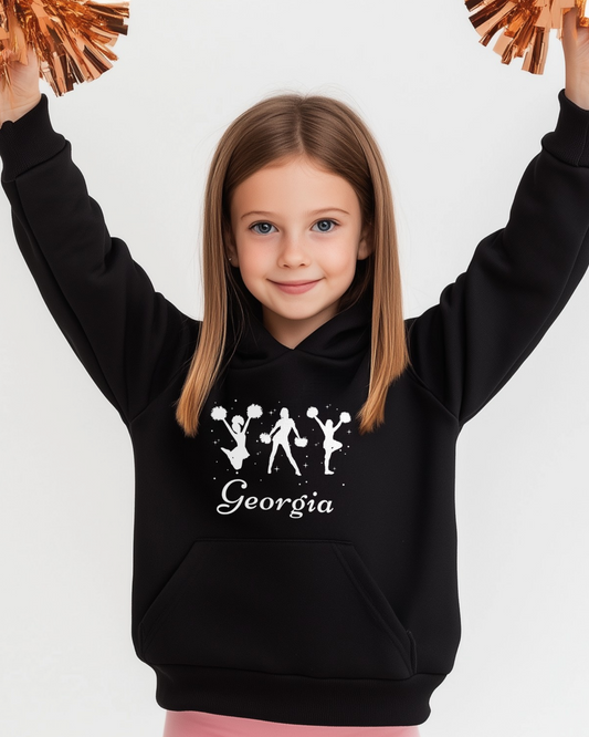 Young girl holding copper-coloured pom poms in the air while wearing a black personalised cheerleading hoodie with white cheerleader silhouettes and the name “Georgia” printed on the front.