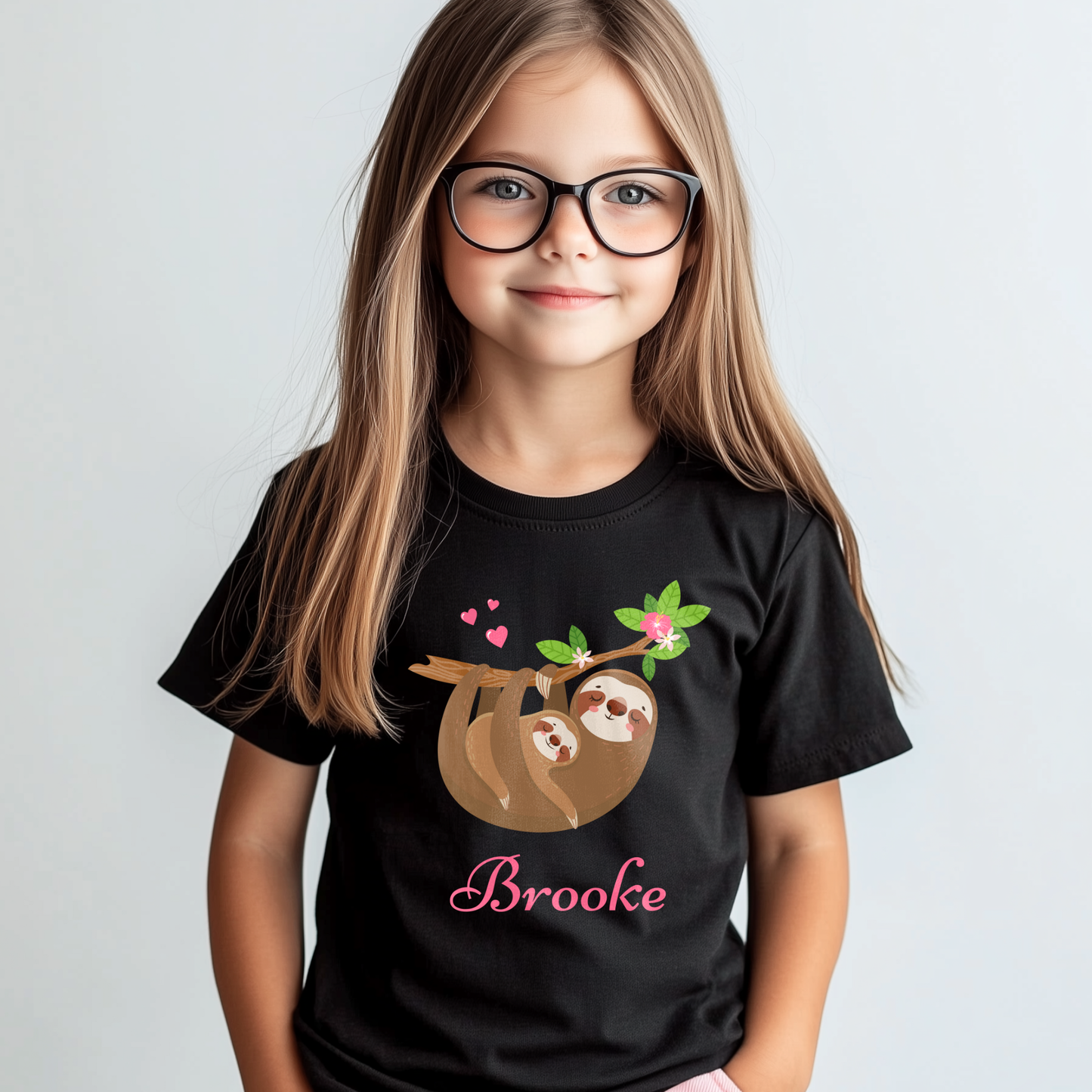 Young girl with long hair and glasses wearing a black T-shirt with a cute mum and baby sloth hanging from a leafy branch with pink hearts and flowers, and the name “Brooke” printed in pink script.