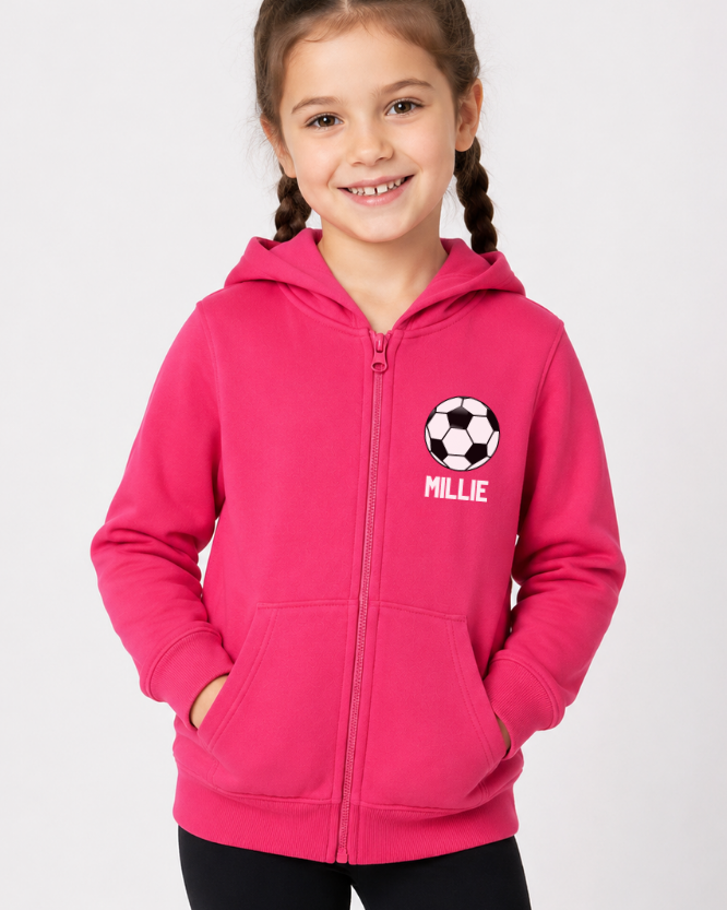 A young girl with braided hair wearing a bright pink zip-up hoodie with a soccer ball graphic and the name 'MILLIE' printed on the chest, posed against a light grey studio background.