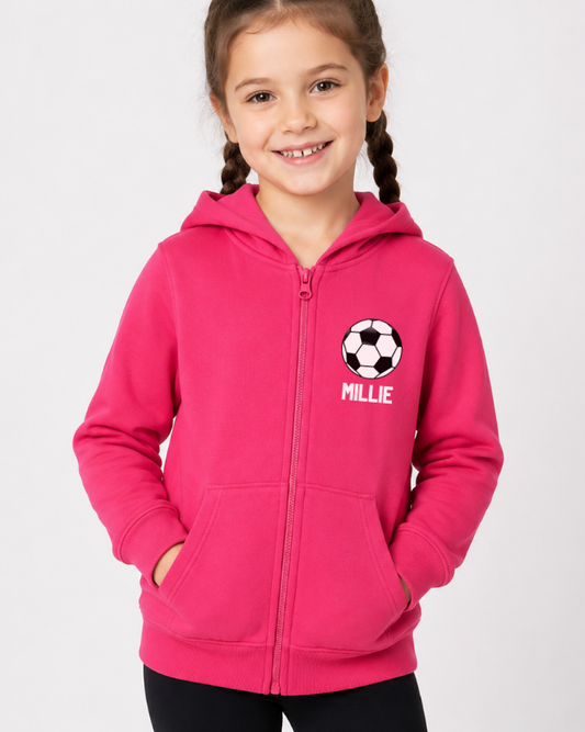 A young girl with braided hair wearing a bright pink zip-up hoodie with a soccer ball graphic and the name 'MILLIE' printed on the chest, posed against a light grey studio background.