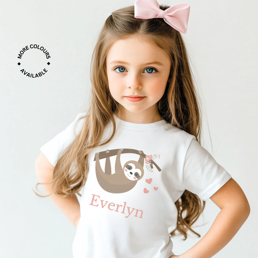 A young girl with long brown hair and blue eyes wears a white t-shirt. The t-shirt features a graphic of a hanging sloth with flowers and hearts, and the name "Everlyn" is printed below the sloth.