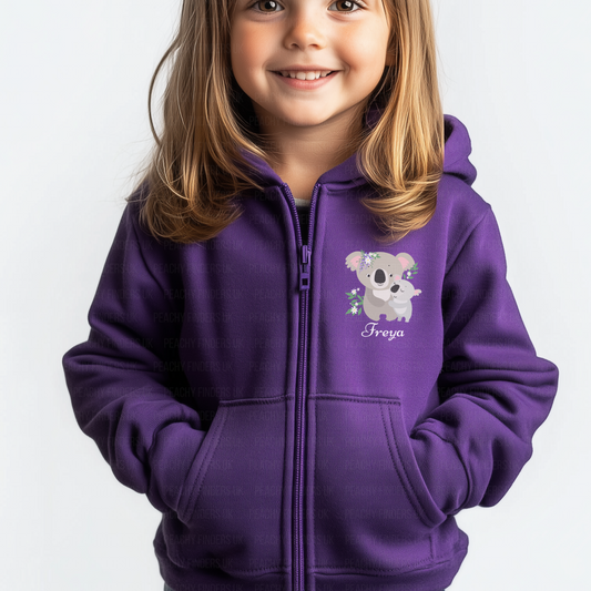 A smiling young child with shoulder-length blonde hair wears a purple full-zip hoodie. On the left chest of the hoodie is a graphic of a grey Mumma and baby koala with lilac floral accents, and below it is the name "Freya" in a light purple script font. The child has their hands in the hoodie's front pockets.