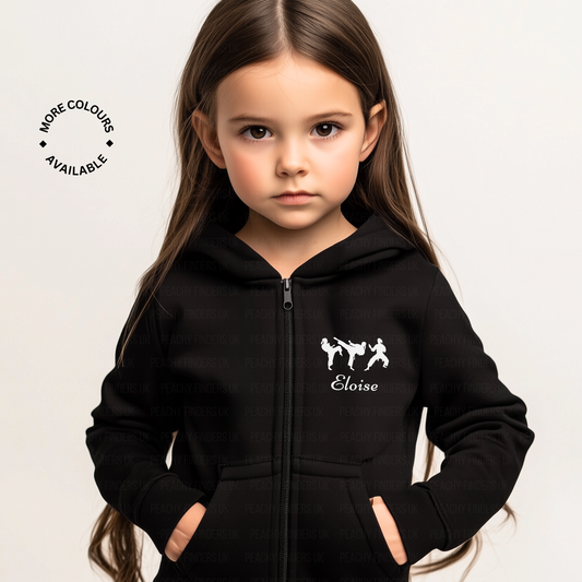 A young girl wearing a black full-zip hoodie, featuring three silhouettes of figures in karate/martial arts poses with the personalised name underneath, her hands in the side pockets of the hoodie.
