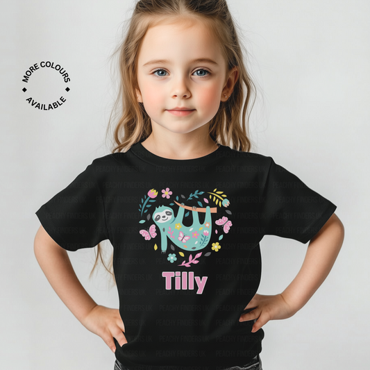 A young girl with long blonde hair wears a black t-shirt. The t-shirt features a graphic of a light blue sloth hanging from a branch, surrounded by pink flowers and hearts. The name "Tilly" is printed below the sloth.
