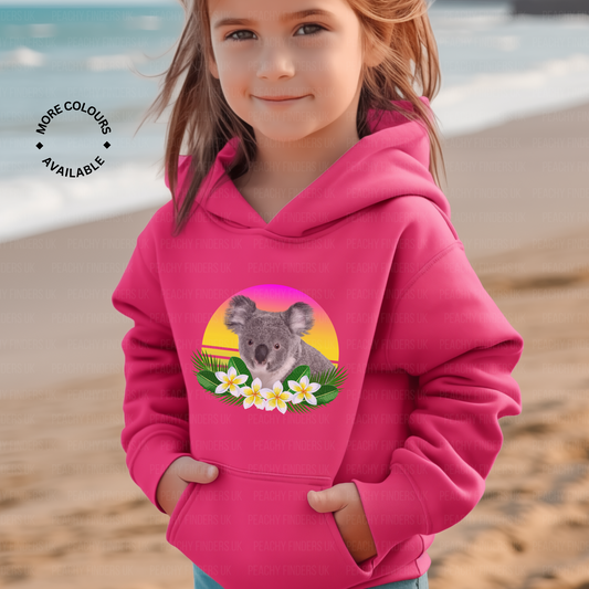 Young girl on the beach modelling a hot pink pullover hoodie featuring a photorealistic koala with tropical flowers against a pink and yellow retro sunset background.