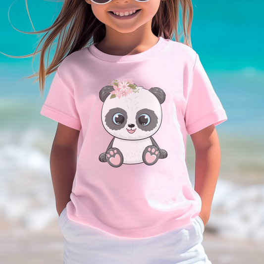A young child smiles, wearing a soft pink t-shirt with a cute panda graphic. The panda has a floral crown on its head. The background is a blurred beach scene with turquoise water and white sand.