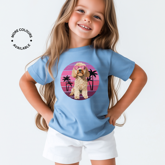 A young girl with long blonde hair models a light blue t-shirt with a graphic of a cockapoo dog wearing pink sunglasses, set against a pink and purple sunset with palm trees.