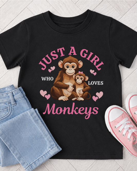 A kids black short-sleeve graphic T-shirt laid flat on a light wood floor, styled with folded light blue jeans and pink sneakers. The shirt features a cartoon mother monkey and baby monkey with pink hearts and the text 'JUST A GIRL WHO LOVES Monkeys' in pink and white.