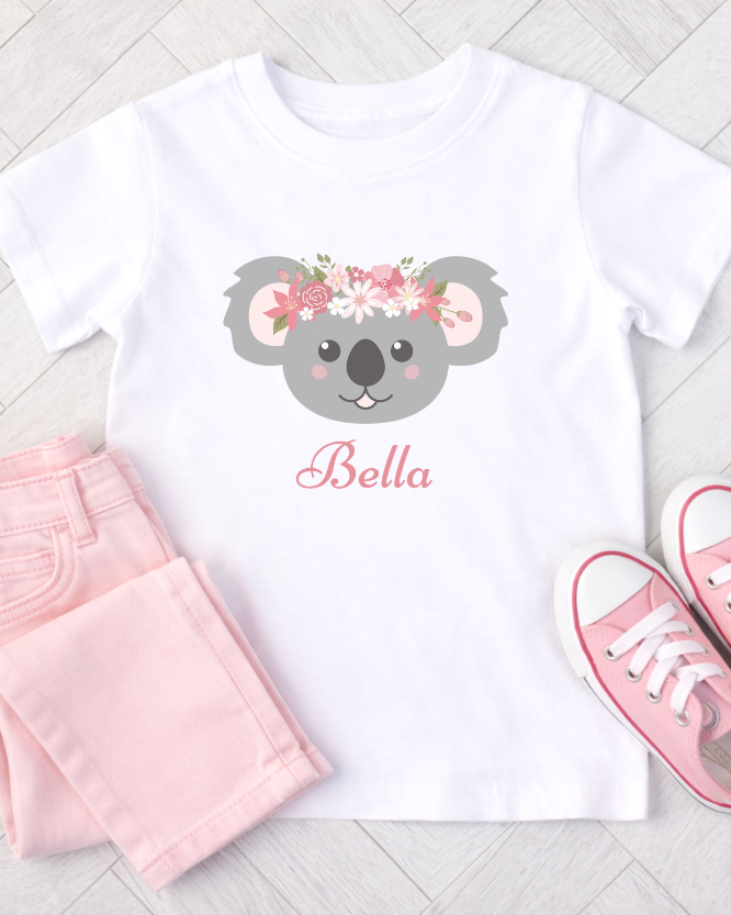 Girls white t-shirt with a koala design and 'Bella' text, paired with pink shorts and shoes on a light wooden floor.