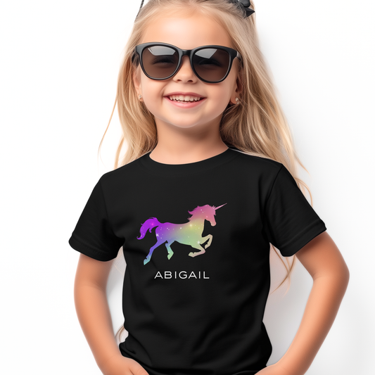 Smiling young girl wearing a black personalised T-shirt featuring a colourful ombre galloping unicorn and the name “Abigail” printed in white below.