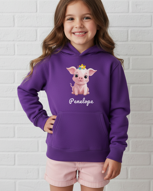 A young girl with long brown hair smiling, wearing a purple pullover hoodie with a cartoon pig wearing a floral gold crown graphic and the name 'Penelope' printed underneath, paired with light pink shorts, standing against a white brick wall.