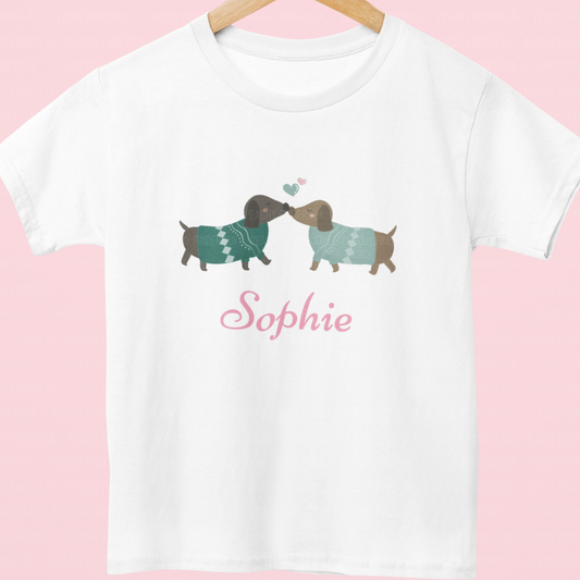Kids white t-shirt with custom name "Sophie" featuring two cute dachshunds in green jumpers kissing, with pastel heart details on a wooden hanger