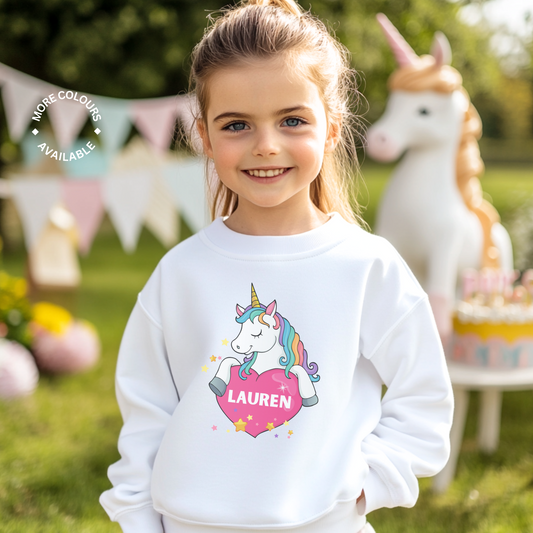 A young girl at a garden party styling a white sweatshirt featuring a colourful unicorn holding a pink heart with the custom name printed inside, surrounded by yellow and pink stars.