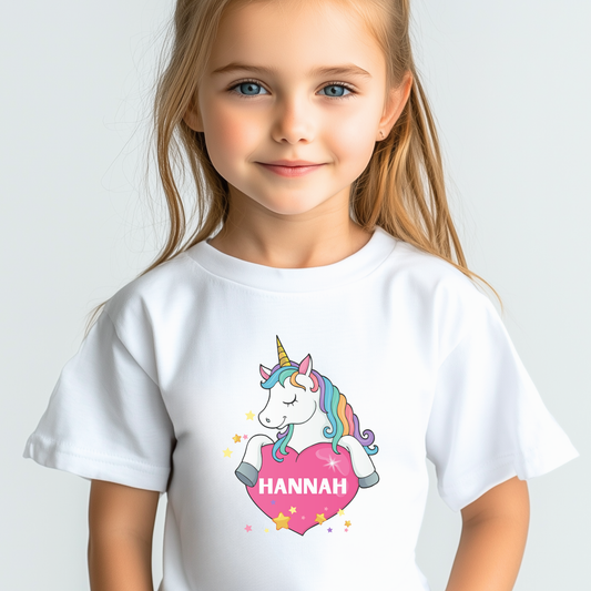 Young girl wearing a white personalised unicorn t-shirt featuring a rainbow-maned unicorn holding a pink heart with the name “Hannah” printed inside, surrounded by yellow and pink stars.