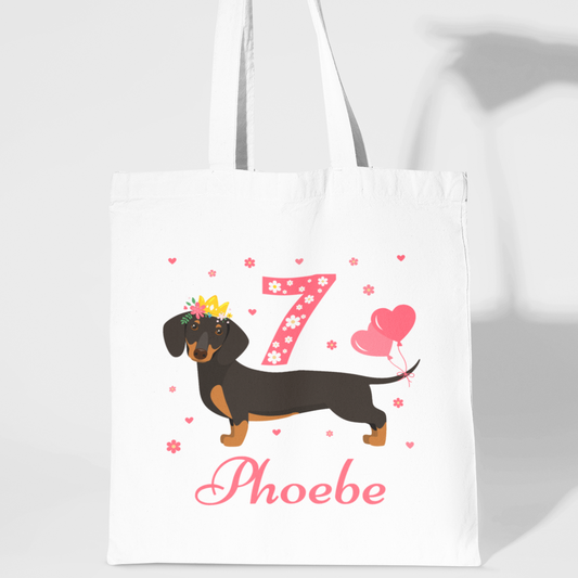 Kids white cotton tote bag with a printed birthday-themed dachshund design, hearts, and the name 'Phoebe' on the front.