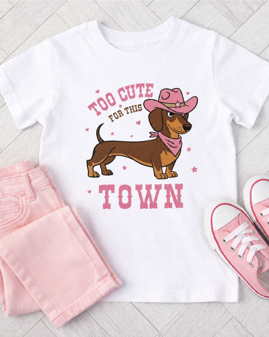 White t-shirt with dachshund graphic and text, pink pants, and shoes on a light wooden floor.