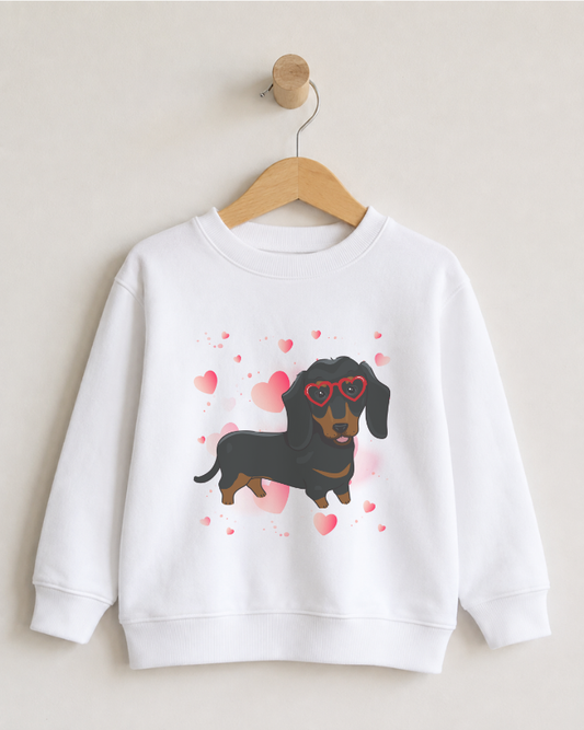 A girls white crewneck sweatshirt hanging on a wooden hanger against a light beige wall. The sweatshirt features a graphic print of a black and tan dachshund wearing red heart-shaped sunglasses, surrounded by red and pink hearts.