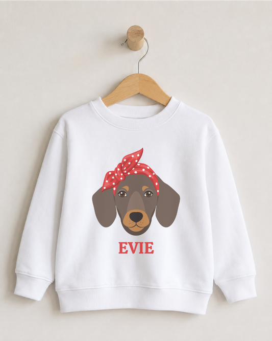 A girls white crewneck sweatshirt hanging on a wooden hanger against a light beige wall. The sweatshirt features a graphic of a dachshund dog wearing a red and white polka-dot bandana, with the name 'EVIE' printed in red below the dog.
