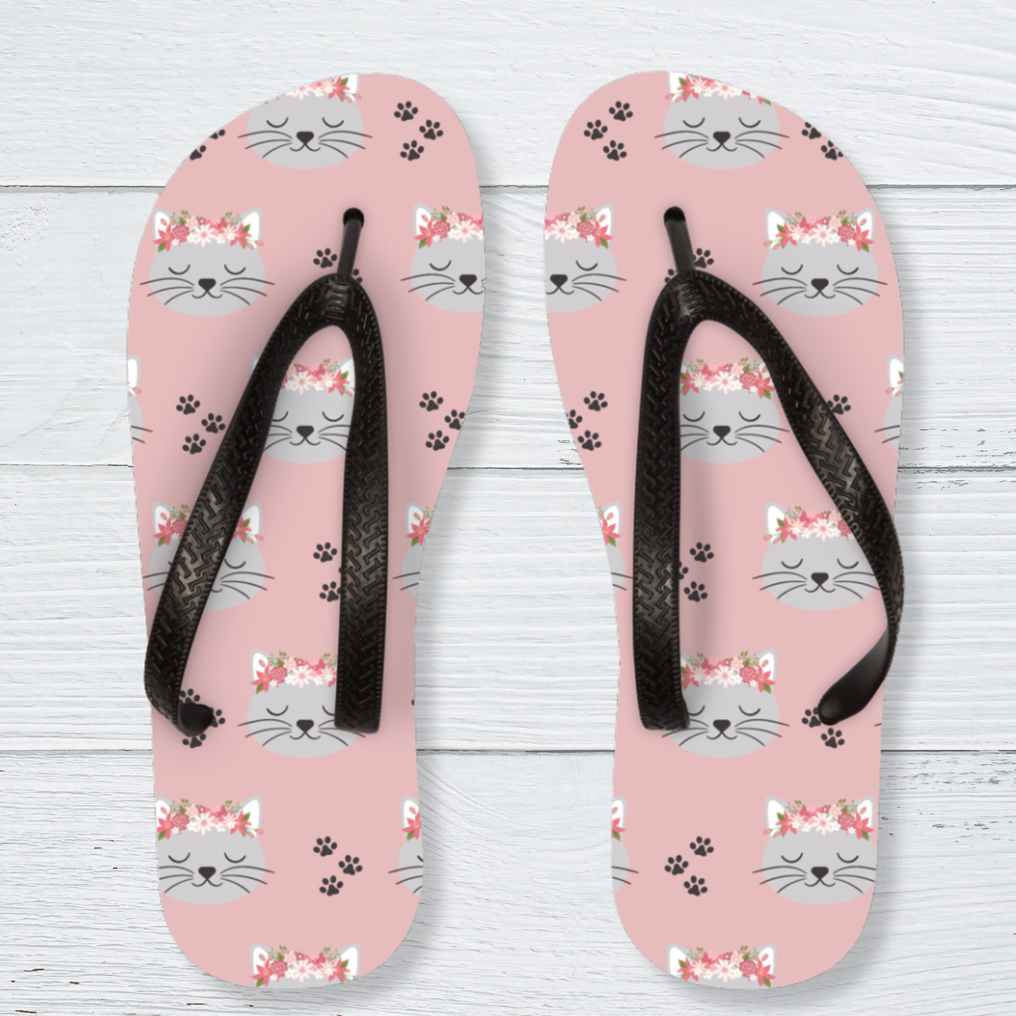 A pair of kids pink flip-flops with a repeating pattern of grey cat faces wearing pink floral crowns, black paw prints, and black woven-look straps, displayed on a white wood plank background.