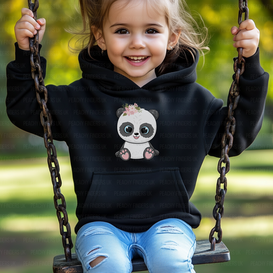 A young child with light brown hair and big brown eyes smiles while sitting on a swing. The child is wearing a black hooded sweatshirt with a cute panda graphic on the front. The panda has a floral crown on its head. The swing hangs from thick, dark chains. The background is blurred green and brown, suggesting a park or garden setting.