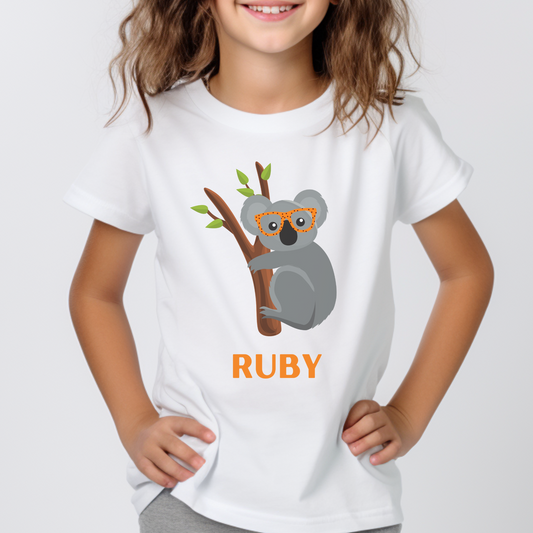 Girl modelling a white short sleeve t-shirt featuring a cute koala wearing orange spotted glasses sitting on a tree branch, personalised with the name Ruby in bold orange text.