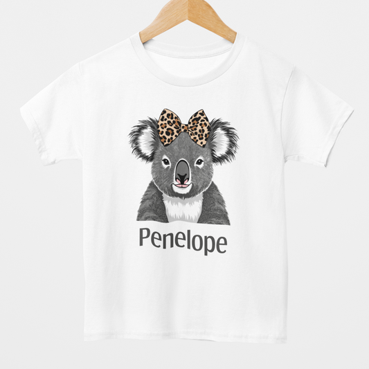 Girls white t-shirt featuring a cute grey koala illustration with a leopard print bow on its head and the name “Penelope” printed underneath in black text. Styled on a wooden hanger.