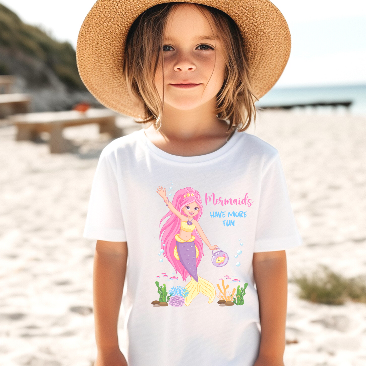 A little girl on the beach wearing a white short sleeved t-shirt with a printed design "Mermaids have more fun" Mermaid with pink hair under the sea.