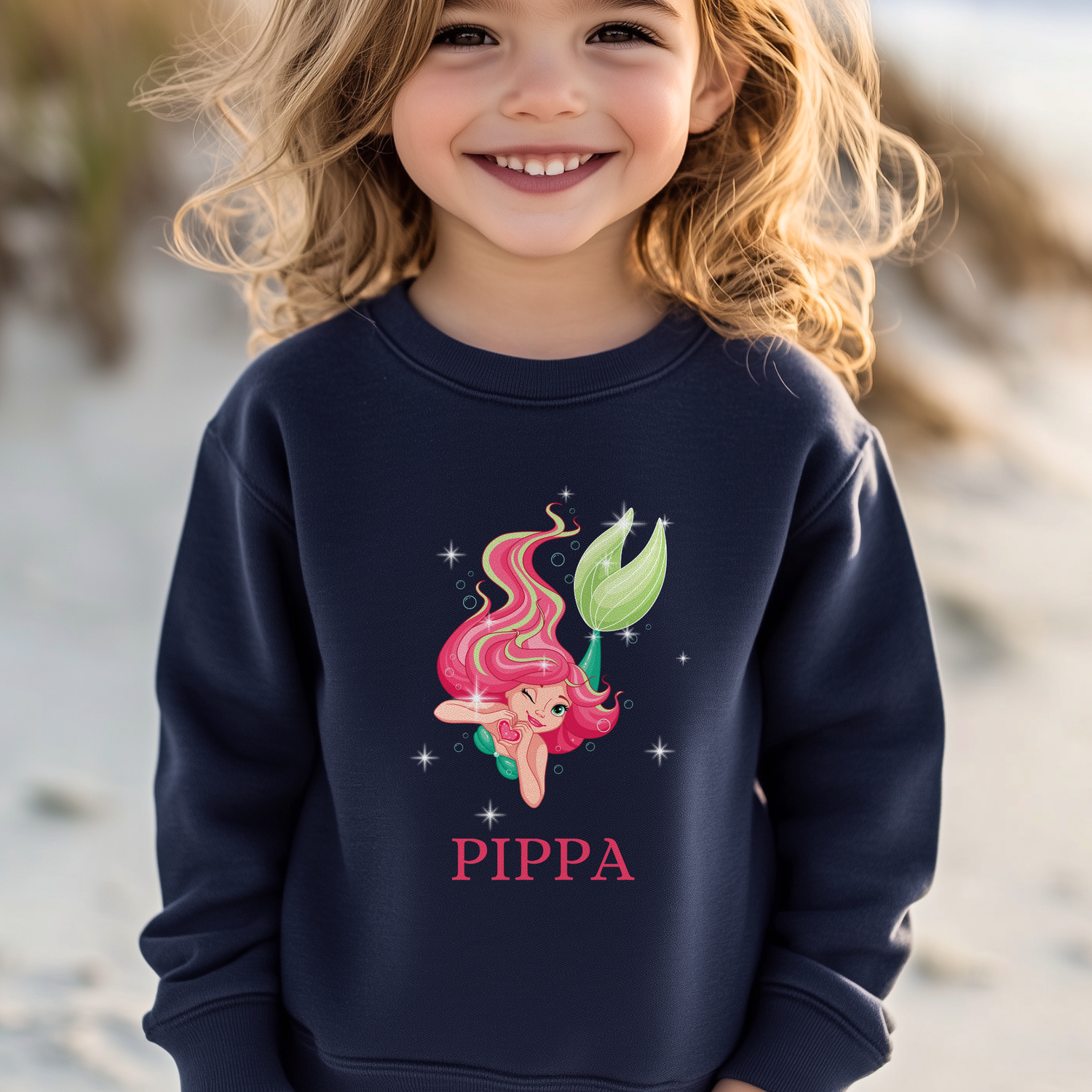 Smiling young girl standing on a beach wearing a navy sweatshirt featuring a pink-haired mermaid surrounded by stars with personalised name “Pippa” printed underneath.