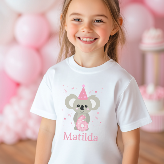 Smiling young girl wearing a white personalised birthday T-shirt featuring a cute grey koala with a pink party hat and number six, surrounded by pink balloons.