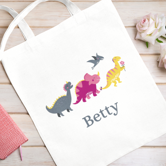 Kids white cotton tote bag with colourful cartoon dinosaur print, featuring a pink triceratops, yellow dino, grey dino, and pterodactyl.