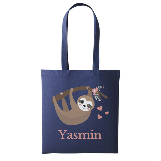 A navy tote bag with a cartoon sloth hanging from a branch surrounded by pink flowers and hearts. The name "Yasmin" is printed below the sloth.