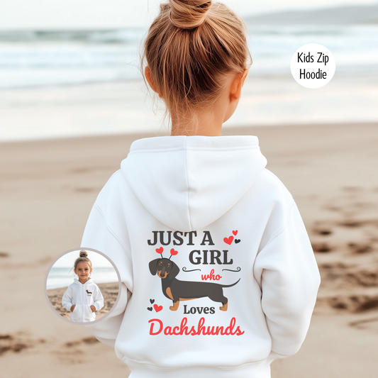 A young girl with blonde hair in a bun faces away from the viewer, wearing a white zip hoodie. The hoodie has the text "JUST A GIRL who Loves Dachshunds" and a graphic of a black and tan dachshund with red hearts. The background is a blurred beach scene with sand and water. An inset image shows the same child from the front, wearing the same hoodie.