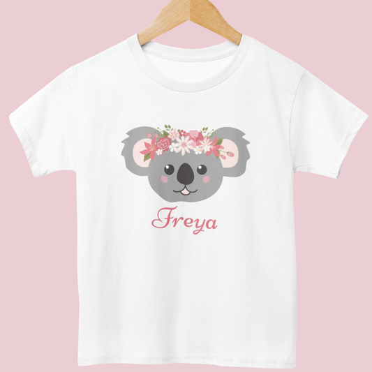 Girls wearing personalised white t-shirt with floral koala graphic and custom name styled on a wooden hanger.