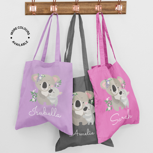 Three personalised cotton tote bags featuring a mother and baby koala with floral designs, printed with the names Isabella, Amelie, and Sarah in pink, grey, and lavender colours.
