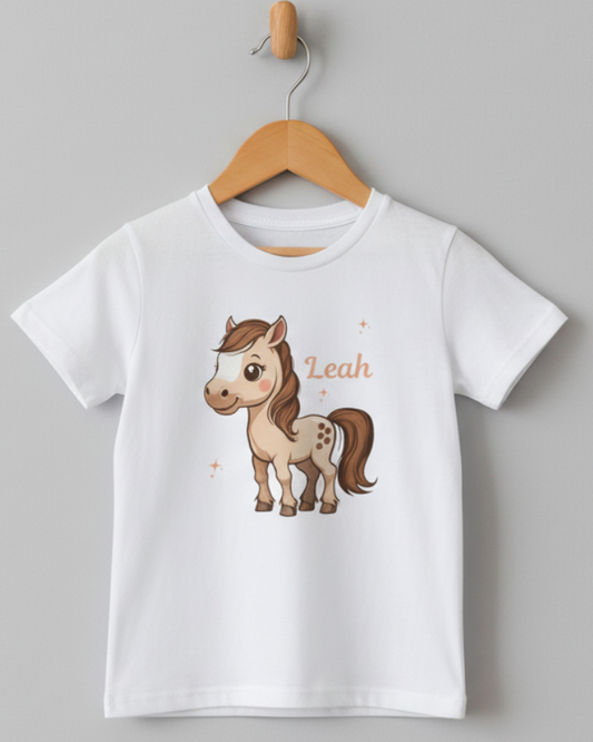 Personalised kids white pony T-shirt with custom name Leah hanging on a wooden hanger against a light grey wall.