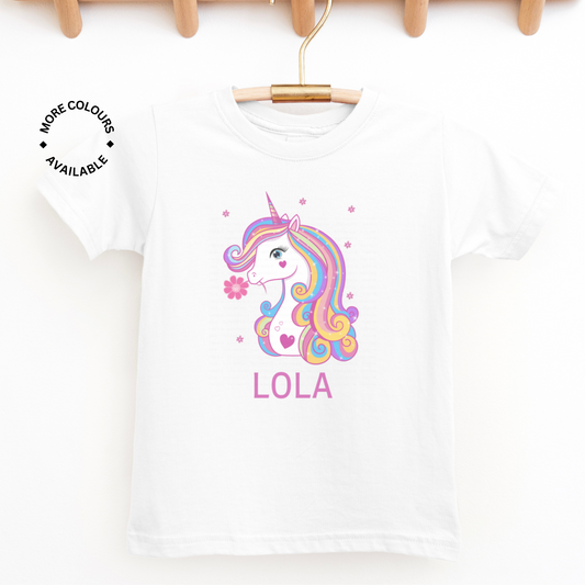 Kids white cotton t-shirt featuring a unicorn in with rainbow hair and pink flowers with custom name, styled on a wooden hanger.