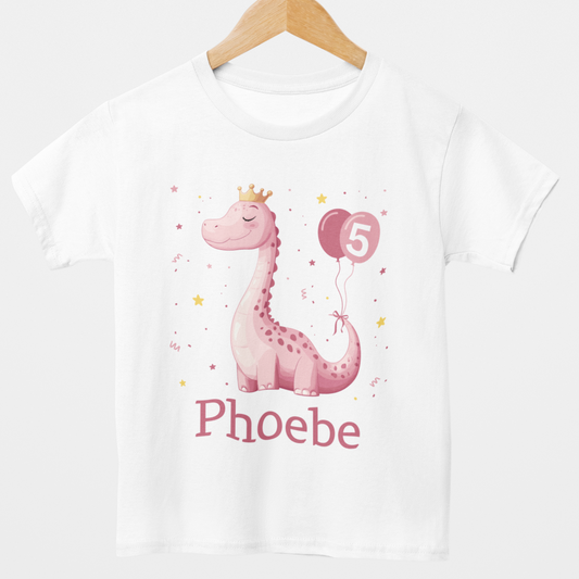 A kids white t-shirt is displayed on a white background. The shirt features a cute, cartoon-style pink dinosaur with darker pink spots along its body and a long neck. The dinosaur is wearing a small golden crown on its head and has its eyes closed with a serene expression. It's holding two pink balloons with the number "3" printed in white on one of them.