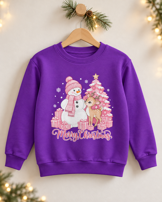 A girls purple crewneck sweatshirt hanging on a wooden hanger against a warm beige wall decorated with pine branches and fairy lights. The sweatshirt features a festive Christmas graphic with a snowman, reindeer, pink Christmas tree, gifts, and 'Merry Christmas' text.