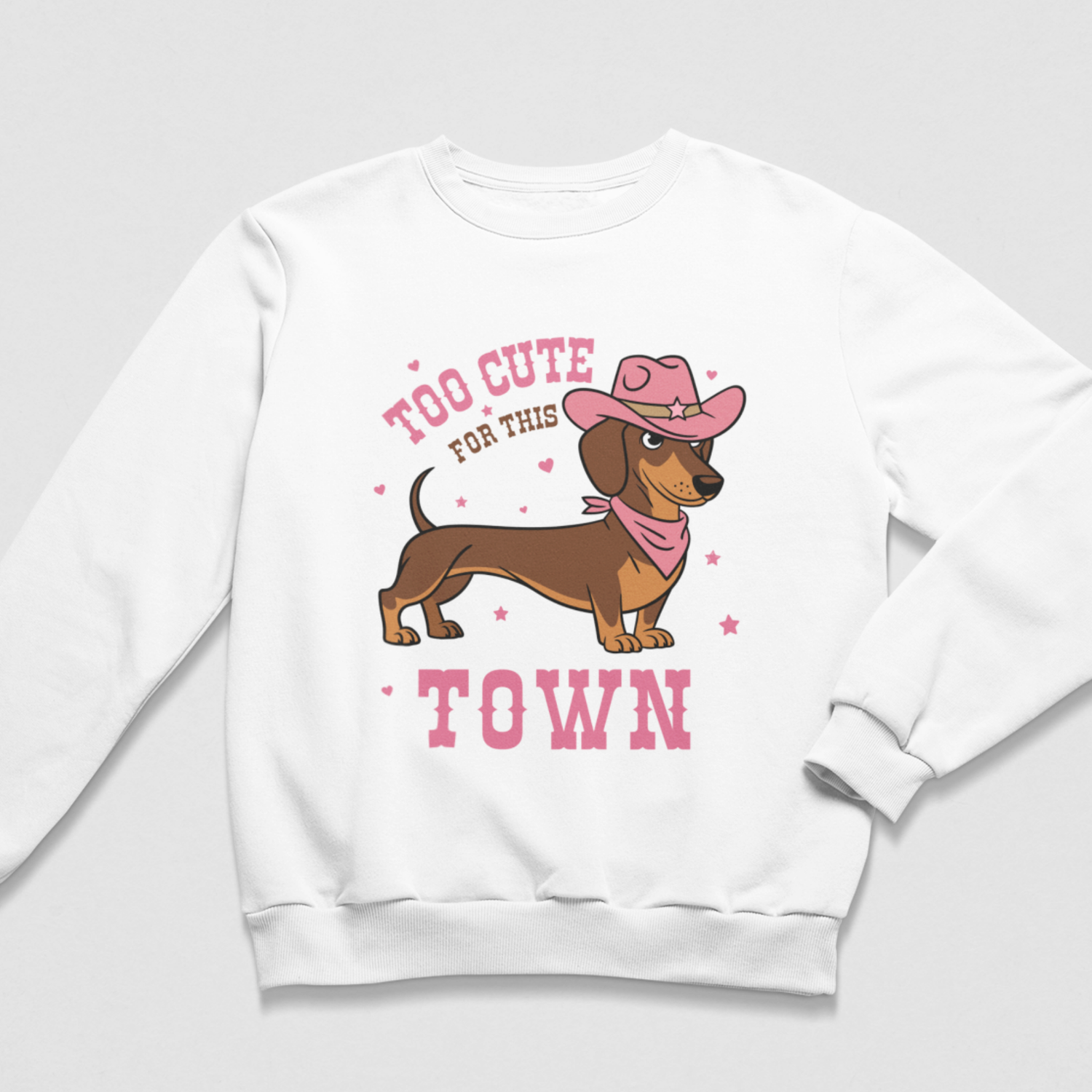 A girls white crewneck sweatshirt featuring a cartoon dachshund wearing a pink cowboy hat and bandana, with the text 'TOO CUTE FOR THIS TOWN' in pink, displayed on a light grey background.